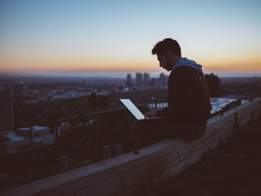 Mann sitzt allein mit Laptop auf einem Dach über der Stadt in der Abenddämmerung